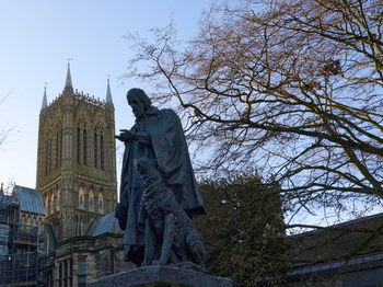 This architectural photograph captures the statue of Lord Tennyson with his dog, prominently displayed in front of the historic Lincoln Cathedral in Lincoln, England. Taken around midday on a bright winter’s day, the image showcases the medieval Gothic architecture of Lincoln Cathedral, a notable landmark in the United Kingdom, with leafless trees providing a seasonal context. The strong daylight and crisp blue sky evoke a clear winter atmosphere, highlighting both the intricate details of the statue and the stonework of the cathedral’s imposing tower. The scene encapsulates a quintessentially English setting, immersing viewers in the heritage and iconic landscape of this part of the United Kingdom.